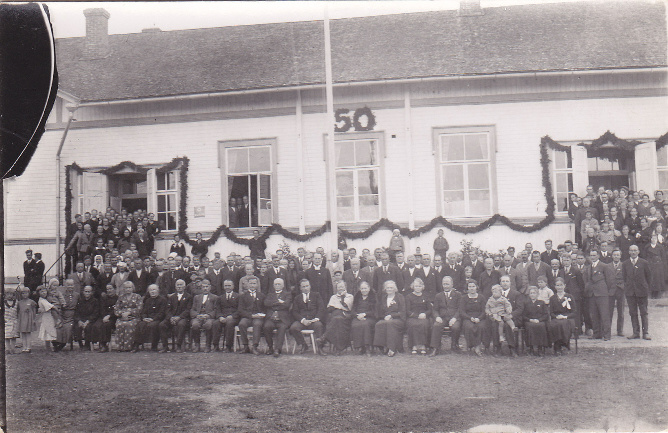 Soanlahden kirkonkylän kansakoulu 50-vuotisjuhlat 1939. Edessä oikealla opettajat Ester Könönen, Olga Kallio, Lauri Ikonen, Maija Ikonen. Lipputangon edessä vasemmalla kirkkoherra Nike Raila. Kanervat köynnöksiin kerättin pappilan mäeltä. Kuva: Seuran arkisto (Siilin).