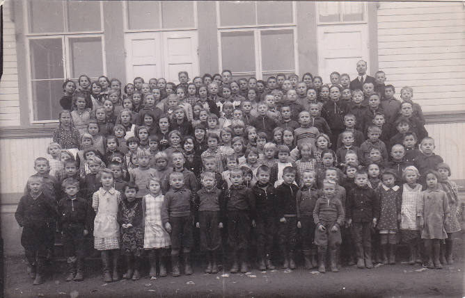 Kirkonkylän kansakoululaisia 1936. Vasemmalla takana opettaja Lauri Ikonen ja vasemmalla oikean oven edessä opettaja Olga Kallio. Kuva: Seuran arkisto (Huurinainen).